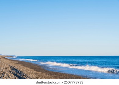 A serene seashore scene featuring a pebble beach with gentle waves lapping against the shore. The vast blue sky stretches above, gradually blending with the horizon where it meets the deep blue ocean. - Powered by Shutterstock