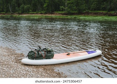 A serene setting by a riverbank shows an inflatable paddleboard with a backpack and paddle, ideal for water sports and ecotourism. The lush forest and rocky shore offer a serene outdoor adventure - Powered by Shutterstock