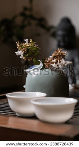 A serene setting on a traditional Chinese tea board: a simple vase with a flower, two white tea bowls in focus, and a Buddha statue subtly standing in the background, creating a Zen-like atmosphere