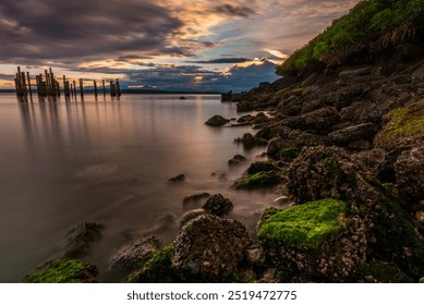 A serene shoreline scene at sunset with a rocky coast covered in green moss, calm waters reflecting the sky, and old wooden pilings extending into the water. The sky is filled with dramatic clouds, pa - Powered by Shutterstock