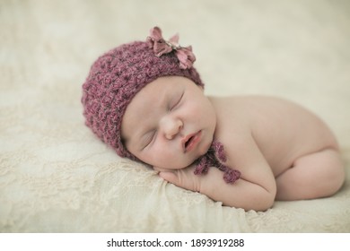 Serene Sleeping Newborn Baby Girl With A Purple Crochet Hat On Her Head Curled Up On A Cream-colored Blanket