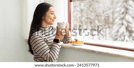 Serene smiling middle eastern millennial woman in striped turtleneck savoring the taste of her morning coffee by a snowy window, her expression peaceful and a croissant on the sill