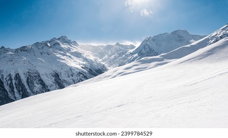 A serene, snow-blanketed mountainous terrain bathed in sunlight, with clear blue skies above and rugged peaks in the distance, suggesting a remote alpine region. - Powered by Shutterstock