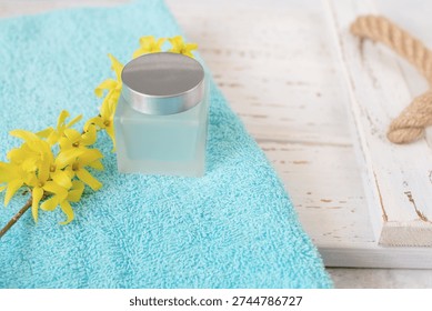 A serene spa setting featuring a soft blue towel, delicate yellow forsythia blossoms, and a frosted glass cosmetic container on a rustic wooden tray