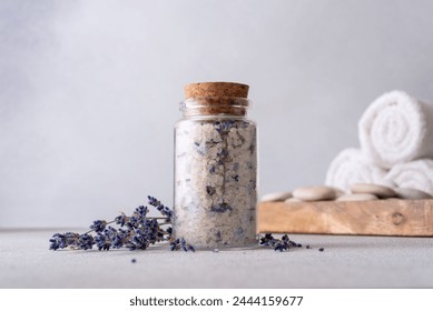 A serene spa setting with a glass jar of aromatic lavender salt sealed with a cork, surrounded by scattered lavender flowers, white towels, and smooth stones against a grey background. - Powered by Shutterstock