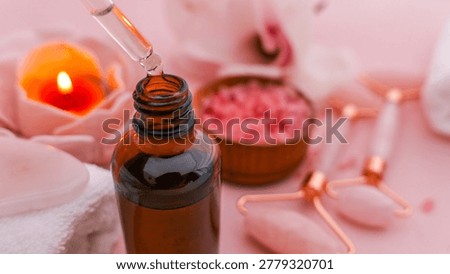 A serene spa setup featuring a dropper dispensing oil into a bottle, alongside a lit candle, pink bath salts, and rose quartz facial rollers