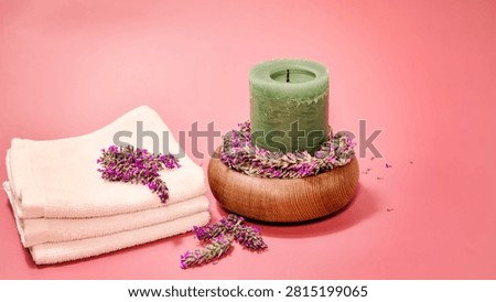 A serene spa setup includes neatly folded white towels and a green pillar candle placed on a wooden bowl adorned with fresh lavender florets, with more lavender scattered nearby on a pink surface.