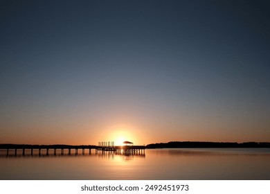 Serene sunset over a calm lake with a silhouette of a wooden pier and gazebo, creating a tranquil and picturesque scene. - Powered by Shutterstock