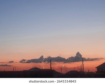 A serene sunset over a quiet town, with silhouettes of rooftops and power lines against a gradient sky of orange, pink, and blue, enhanced by scattered clouds.
