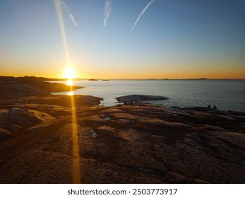 A serene sunset over a rocky coastline with calm waters reflecting the golden sunlight, with distant islands on the horizon and a clear sky streaked with faint contrails. - Powered by Shutterstock