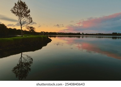 Serene sunset over a tranquil lake with a lone tree reflecting on the water's surface, surrounded by lush greenery and a colorful sky. - Powered by Shutterstock