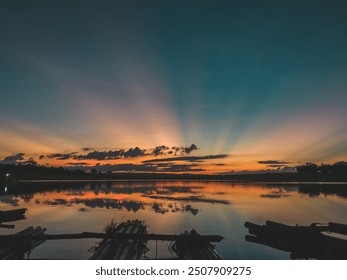 A serene sunset reflecting on calm water, where the sky is painted with shades of blue, orange, and yellow. Dark clouds and silhouettes of trees and rafts add depth to the peaceful scene - Powered by Shutterstock