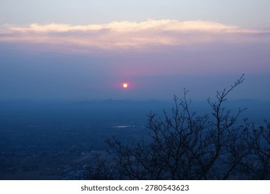 A serene sunset view from an elevated viewpoint (Yelagiri- hills in Tamil Nadu) featuring a glowing orange sun in a hazy purple sky, framed by silhouetted bare tree branches in the foreground