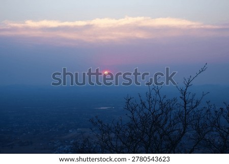 A serene sunset view from an elevated viewpoint (Yelagiri- hills in Tamil Nadu) featuring a glowing orange sun in a hazy purple sky, framed by silhouetted bare tree branches in the foreground