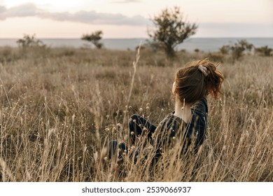 serene sunset woman sitting in tall grass gazes at horizon over ocean at dusk - Powered by Shutterstock