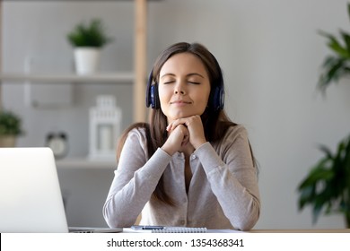 Serene tranquil young woman closed eyes wearing headphones sitting at desk use laptop listening online music, enjoy e-learning easy foreign language. Self-education modern wireless tech usage concept