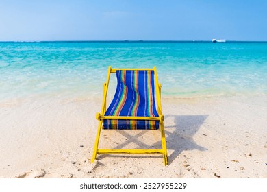 A serene tropical beach scene featuring a vibrant yellow and blue beach chair on pristine white sand, overlooking clear turquoise waters and lush green hills under a clear blue sky. - Powered by Shutterstock