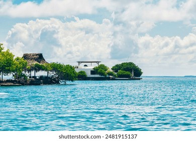 Serene tropical coastline featuring a thatched roof hut, lush greenery, and calm blue waters under a bright sky with fluffy clouds. - Powered by Shutterstock