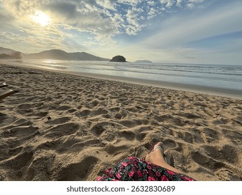 A serene view of a beach at dawn, capturing a person's feet resting in the light sand. The soft sunlight rising on the horizon reflects on the calm water, with mountains in the background. 