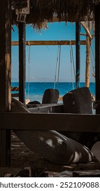 A serene view of a beachfront seating area shaded by a rustic wooden structure with the ocean and clear sky in the background. The relaxed ambiance and natural setting evoke a peaceful, laid-back atmo