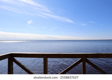 A serene view of the calm river from a wooden dock, with the horizon stretching far into the distance, framed by the wooden railing, and bright blue skies reflecting on the water. - Powered by Shutterstock