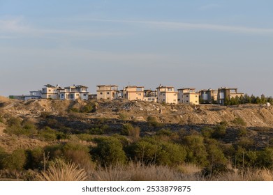 A serene view captures a hillside village bathed in warm sunset light, showcasing modern homes harmoniously set against a backdrop of gentle rolling hills and greenery. - Powered by Shutterstock