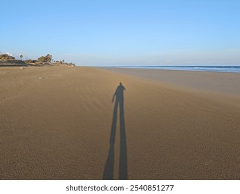 A serene view of an empty beach with a long shadow cast by the photographer, stretching across the sandy shore towards the calm ocean under a clear blue sky, evoking solitude and tranquility