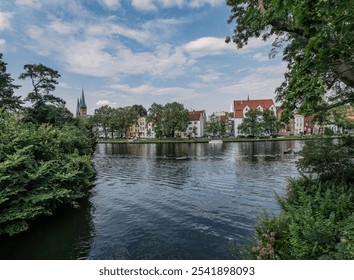 A serene view of historic European architecture by a calm river, surrounded by lush greenery. The picture captures nature's beauty and urban charm under a vibrant sky. - Powered by Shutterstock