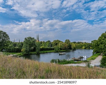 A serene view of historic European architecture by a calm river, surrounded by lush greenery. The picture captures nature's beauty and urban charm under a vibrant sky. - Powered by Shutterstock