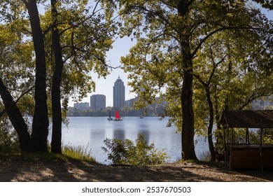 A serene view of a lake framed by trees, with red sailboats and a cityscape in the background under a clear sky - Powered by Shutterstock