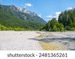 A serene view of a parched riverbed with patches of water, surrounded by lush forests and the towering peaks of the Julian Alps in Triglav National Park. Jasna near Kranjska Gora, Slovenia