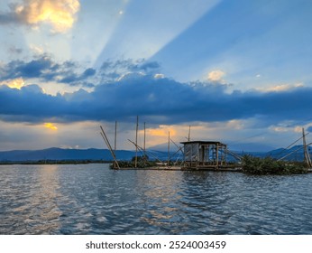 Serene view of Rawa Pening lake at sunset. Wooden hut and fishing nets (Branjang) stand on stilts in water. Sky glows in dramatic orange and blue, with streaks of sunlight piercing through clouds. - Powered by Shutterstock