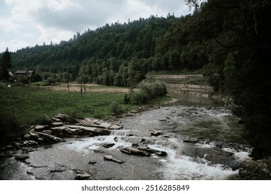 A serene view of a river winding through lush greenery, with rocky banks and a distant rustic house under an overcast sky. - Powered by Shutterstock