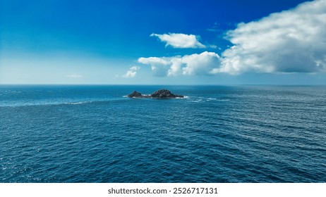 A serene view of a small rocky island surrounded by calm blue ocean waters under a clear sky with fluffy white clouds. At Land's End, Cornwall, UK - Powered by Shutterstock