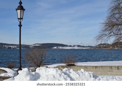 Serene view of Tegernsee Lake in winter, framed by snow-covered banks and a classic lamppost, with a clear blue sky and scenic hills in the background - Powered by Shutterstock