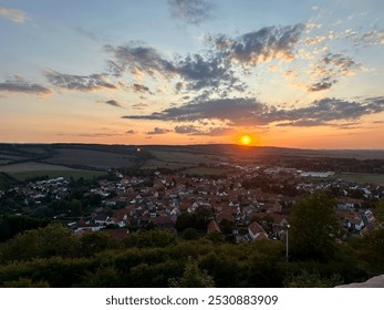 A serene village landscape captured at sunset, with the sun setting just above the horizon, casting warm hues of orange, pink, and purple across the sky. The village below with red-roofed houses. - Powered by Shutterstock