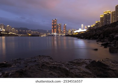 A serene waterfront scene with illuminated, uniquely shaped skyscrapers against a dusky sky reflecting on calm waters near rocky shores. - Powered by Shutterstock