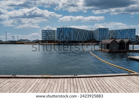A serene waterfront with a wooden pier, metal cleat, and orange life ring in Copenhagen, featuring modern buildings, a yellow barrier line, and floating structures under a partly cloudy sky.