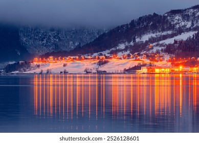 A serene winter evening reveals a charming village with glowing lights reflecting on a calm fjord, surrounded by snow-laden mountains. - Powered by Shutterstock