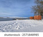 Serene winter landscape featuring a snow-covered lakeshore with a wooden pier extending into the calm water. Two wooden structures stand near leafless trees under a clear blue sky