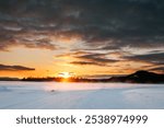 Serene winter landscape at sunset with dramatic clouds and snow-covered ground, creating a peaceful scene of nature’s beauty and tranquility