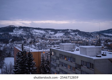 A serene winter view of a European town at dusk. Snow-covered apartment buildings with warm glowing windows, hills, and trees create a cozy atmosphere under an overcast sky. - Powered by Shutterstock