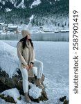 Serene Winter Wonderland: Young Woman in Cozy Cream Outfit Sitting on Snowy Rock Overlooking Frozen Eibsee Lake and Zugspitze Mountain