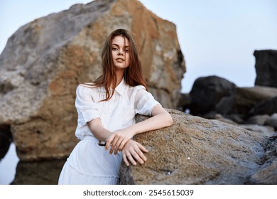 Serene woman in elegant white dress sitting on rocky shore with hands on hips, gazing out at ocean - Powered by Shutterstock