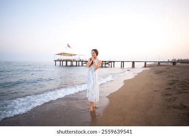 Serene Woman in Flowing Dress Strolling on Sandy Beach by Wooden Pier at Sunset  - Powered by Shutterstock