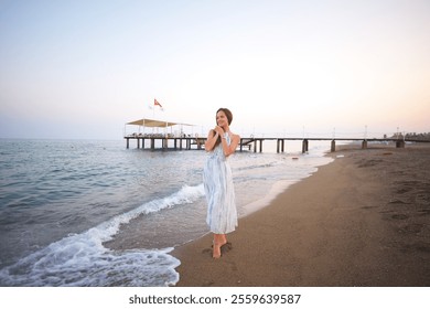 Serene Woman in Flowing Dress Strolling on Sandy Beach by Wooden Pier at Sunset  - Powered by Shutterstock