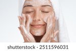 A serene woman with a towel wrapped around her head gently massages foaming cleanser into her skin, enjoying a peaceful moment during her skincare routine. Ideal for promoting beauty, self-care, and