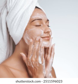 A serene woman with a towel wrapped around her head gently applies a foaming cleanser to her face, enjoying a refreshing skincare routine. Ideal for promoting beauty, self-care, and healthy skin - Powered by Shutterstock