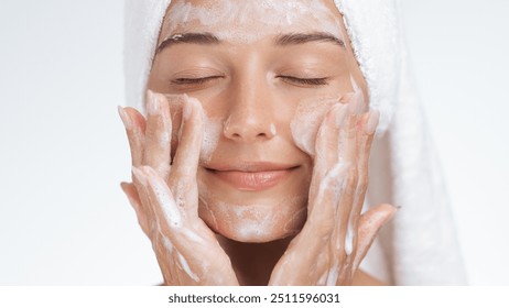 A serene woman with a towel wrapped around her head gently massages foaming cleanser into her skin, enjoying a peaceful moment during her skincare routine. Ideal for promoting beauty, self-care, and - Powered by Shutterstock