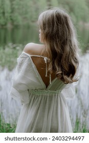 A Serene Woman in a White Dress by a Tranquil Body of Water posing. Female or bride in wedding long transparent dress in forest. Wild beach near laker, pond or sea. Summer vibes. Unrecognizable girl.